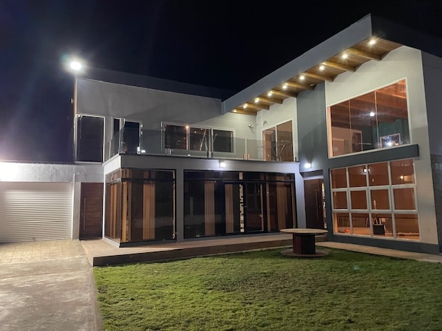 The modern villa is illuminated at night, highlighting large glass windows and an expansive terrace. The outdoor area features a manicured lawn and a circular seating arrangement, creating a comfortable space for relaxation. The sleek architectural design emphasizes clean lines and an inviting ambiance.
