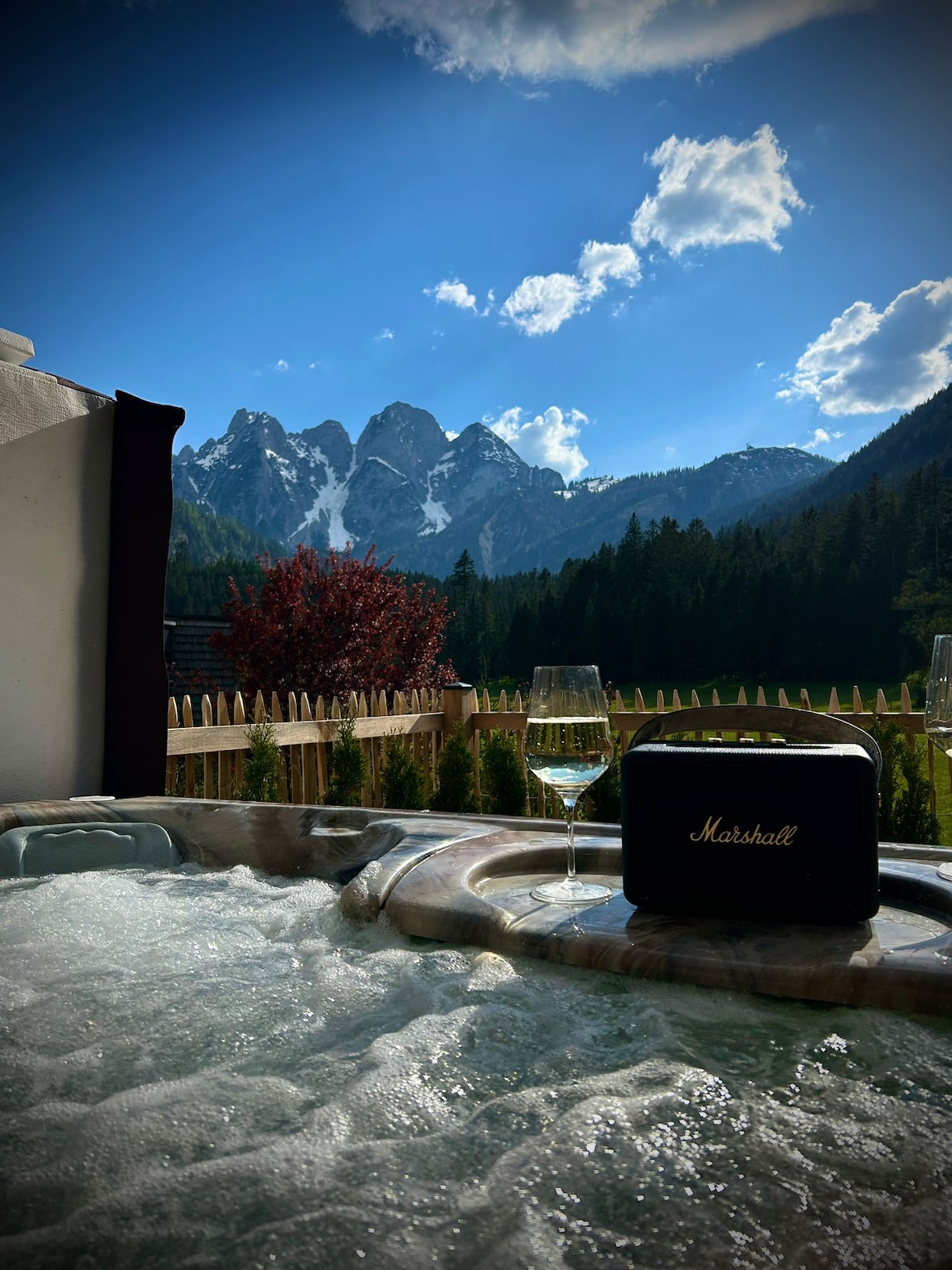 A hot tub is positioned outdoors, surrounded by scenic mountains and lush greenery. A glass of wine rests on the edge of the tub, alongside a portable speaker. The sky is clear, with sunlight illuminating the mountainous backdrop.