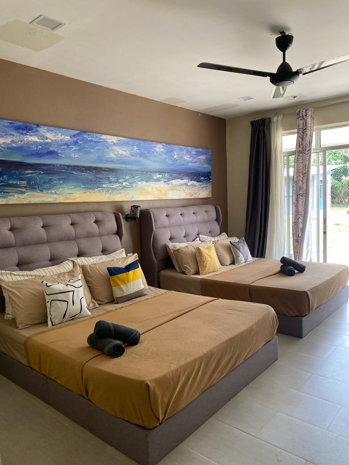 A well-furnished guest room features two spacious beds, each topped with neutral-toned bedding and decorative pillows. A large artwork depicting a coastal scene hangs above the beds, while natural light streams in through sliding glass doors leading to an outdoor area.