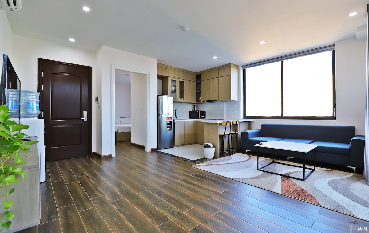 A modern living space features a comfortable sofa and a coffee table situated on a patterned rug. A wall of windows allows natural light to brighten the area. A kitchen is visible in the background, equipped with contemporary appliances and cabinetry.