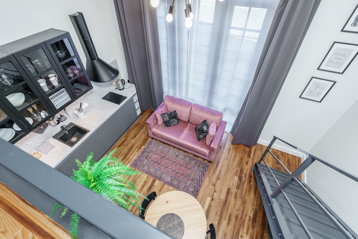 The loft's layout showcases a stylish kitchen with dark cabinetry and light countertops, complemented by a round dining table. A pink sofa sits atop a patterned rug, with large windows dressed in sheer curtains allowing natural light to fill the space.