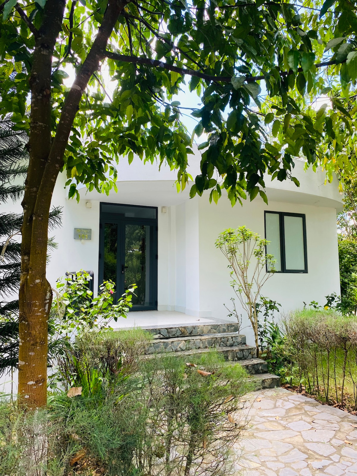 A modern villa entrance is framed by lush greenery and tall trees. Stone steps lead up to a white exterior with large glass doors. The surrounding area features well-maintained shrubs and plants, creating a serene and inviting atmosphere.