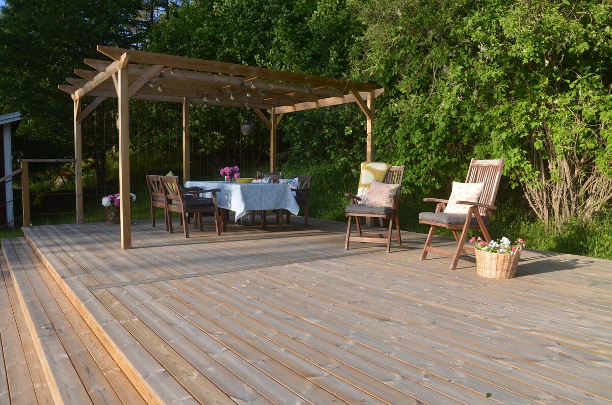 A spacious wooden deck features a covered dining area with a table set for outdoor meals. Comfortable seating is arranged for relaxation, surrounded by lush greenery. Brightly colored flowers are placed in a basket nearby, enhancing the inviting atmosphere.