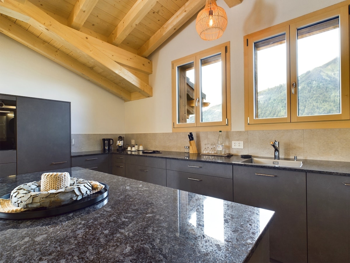 A modern kitchen features a spacious island topped with polished granite. Wooden beams accentuate the ceiling, and large windows display views of the surrounding mountains. Essential appliances, including a coffee maker, are neatly arranged on the counter, while utensils and glassware are organized nearby.