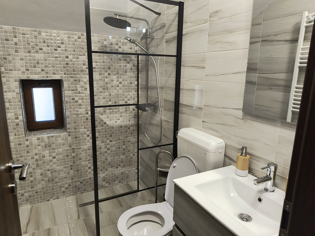 A modern bathroom is presented with a glass shower enclosure and contemporary fixtures. The tiled walls feature a mosaic pattern, while a small window allows natural light. A vessel sink rests atop a sleek vanity, complemented by a white toilet. Neutral tones dominate the space.