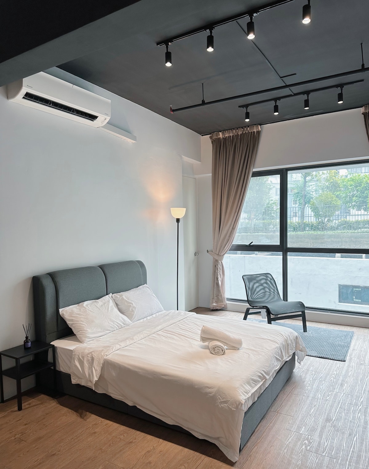 A studio bedroom features a spacious bed with white linens neatly arranged. Beside it, a small bedside table provides additional surface space. Natural light filters through a large window, partially covered by elegant curtains. A chair is positioned in the corner, contributing to the cozy layout.