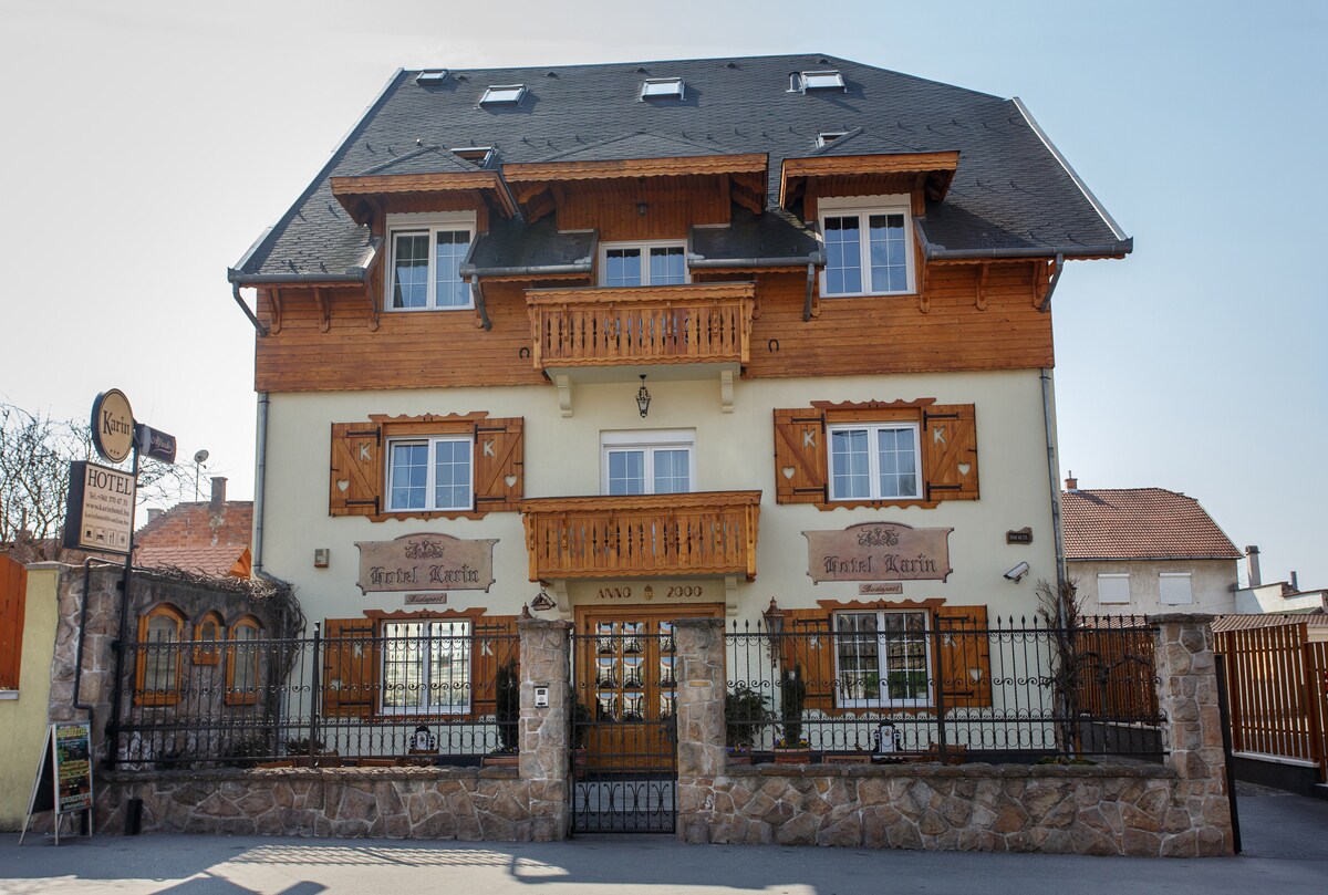 The exterior of a charming hotel is shown, featuring a combination of wood and stone elements. Large windows and a prominent balcony are visible, complemented by decorative carvings. A wrought-iron fence surrounds the property, providing a warm and inviting entrance.