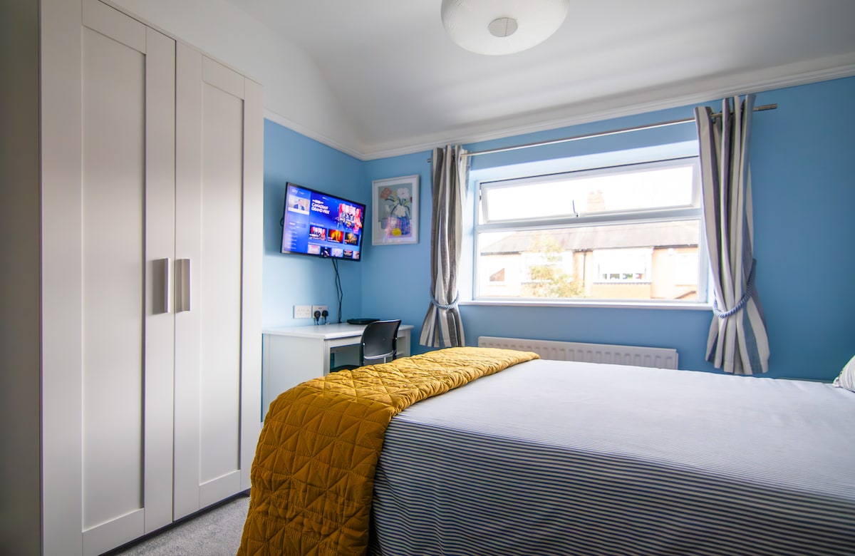 A serene bedroom features a spacious bed with a striking yellow quilt and blue walls. A desk with a chair is positioned next to a wall-mounted television, and large windows allow natural light to fill the space, complemented by striped curtains.