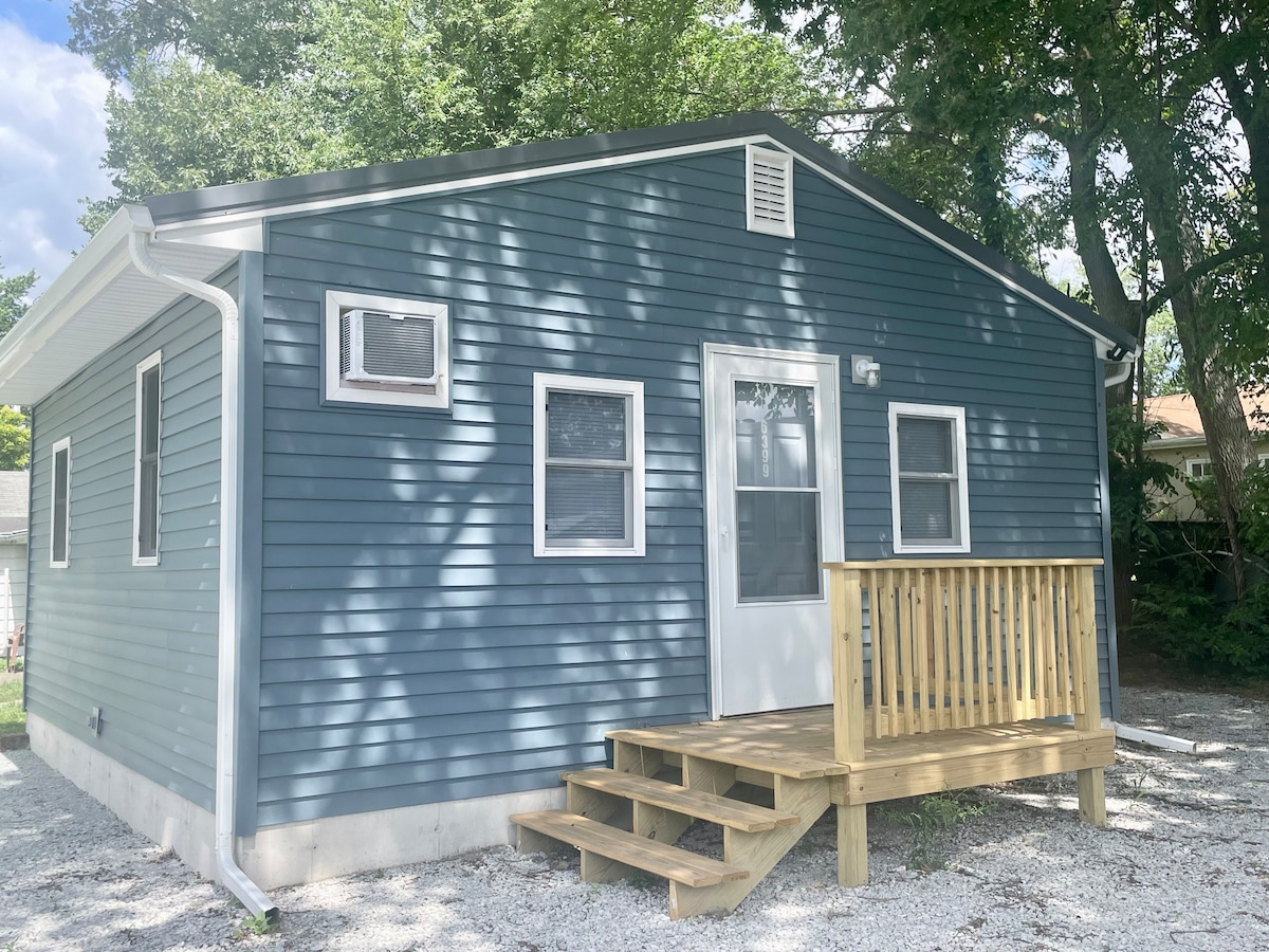 A single-story cottage features a blue exterior with white trim. Steps lead up to a wooden porch with railing, positioned under a canopy of trees. Multiple windows are visible, allowing natural light to enhance the inviting atmosphere.