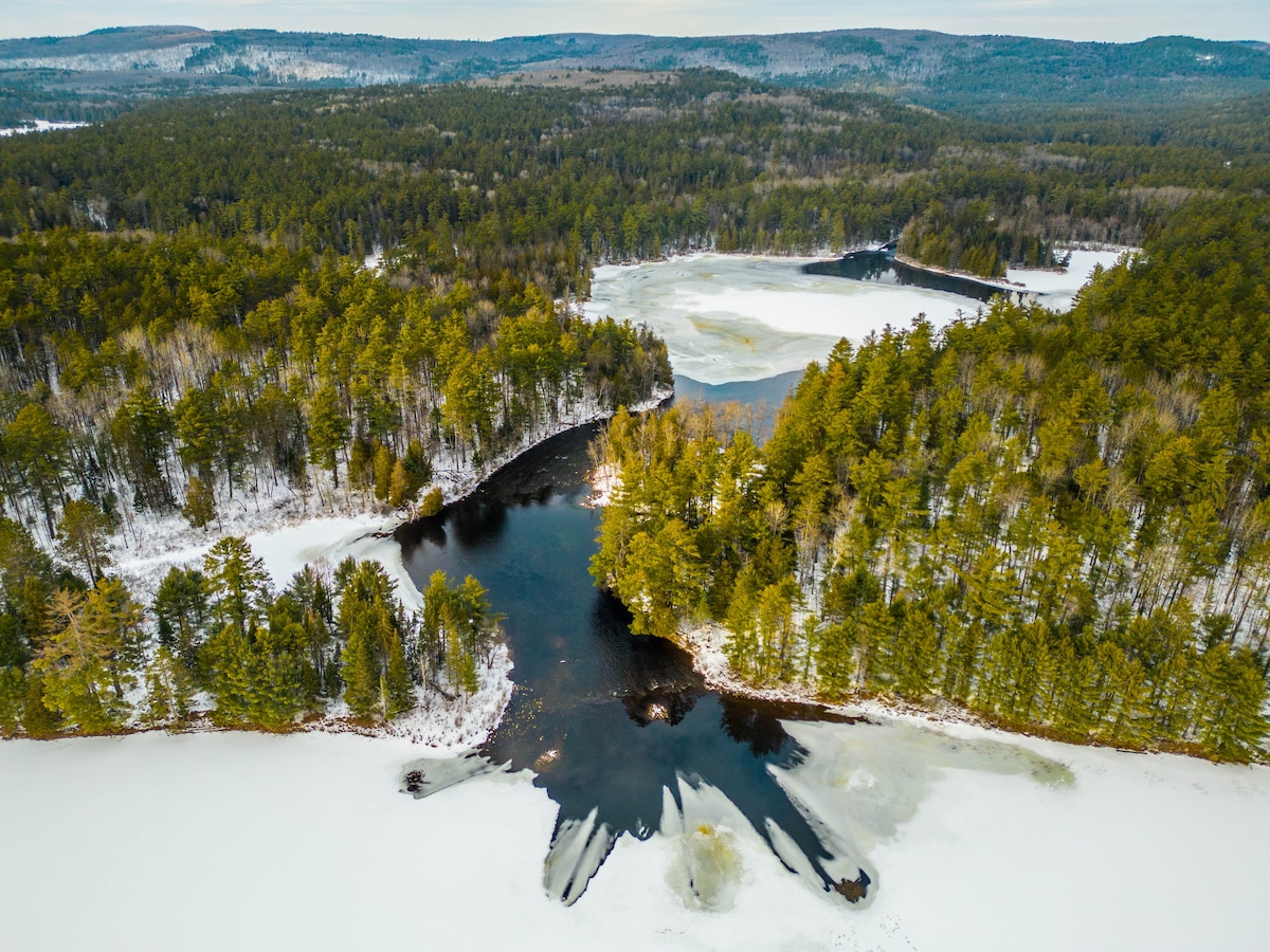An aerial view captures the serene landscape of Pimisi Bay, where dark water flows through patches of snow-covered ice. Surrounding evergreen trees and distant hills harmonize with the natural beauty of the winter scenery, offering a tranquil escape in a forested setting.
