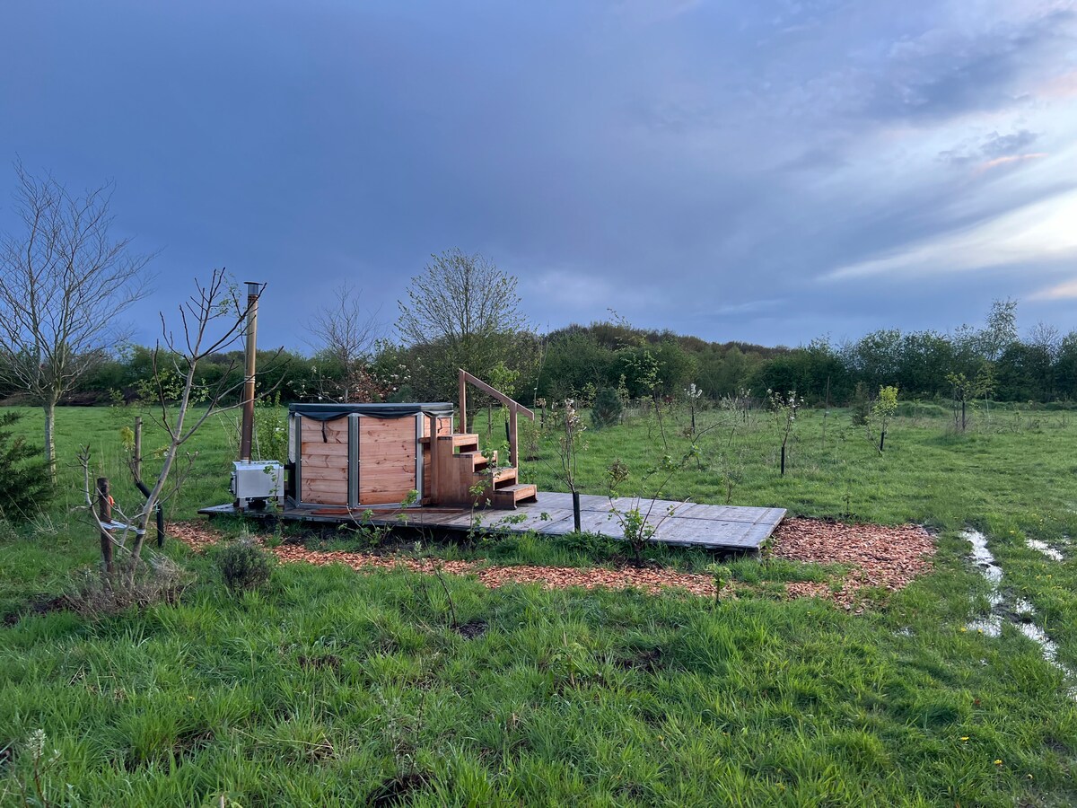 A wooden hot tub is nestled on a raised wooden deck, surrounded by a grassy area. Steps lead up to the tub, while young trees and a lush landscape are visible in the background under a cloudy sky.