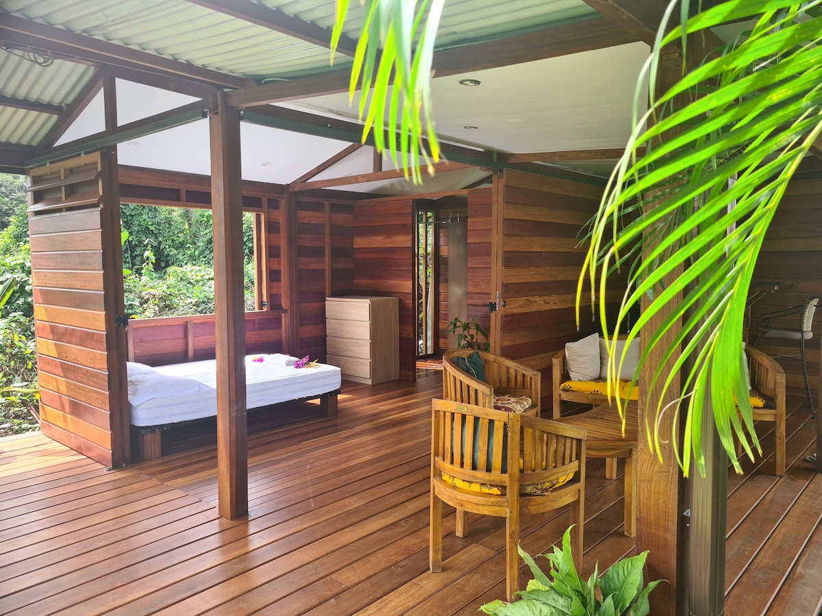 A wooden structure showcases an open living area connected to a bedroom, featuring natural wood walls. A spacious bed is placed beside a dresser. Comfortable seating is arranged in the living space, surrounded by tropical greenery and sunlight filtering through the leaves.