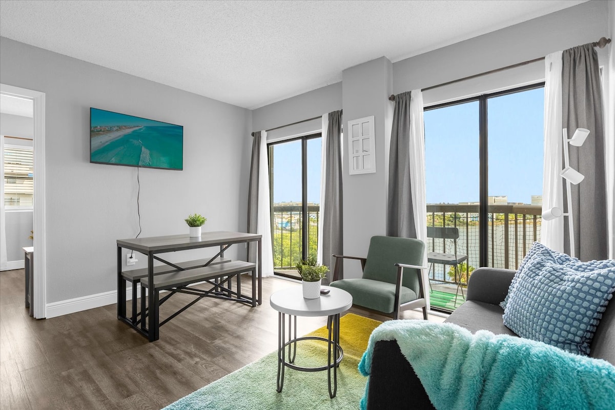 The living area features large windows that provide ample natural light and views of the surroundings. A smart TV is mounted on the wall opposite a modern sofa with a textured blanket draped over it. A round table and comfortable chair complete the cozy atmosphere.