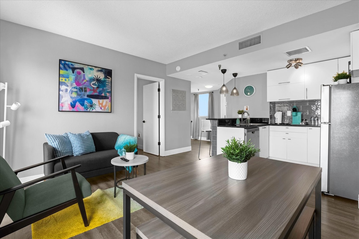 An open-concept living area showcases modern furniture, including a dark couch and a round coffee table with a small plant. Bright artwork adorns the wall, while natural light filters through from a nearby window. A spacious kitchen with sleek appliances is visible in the background.