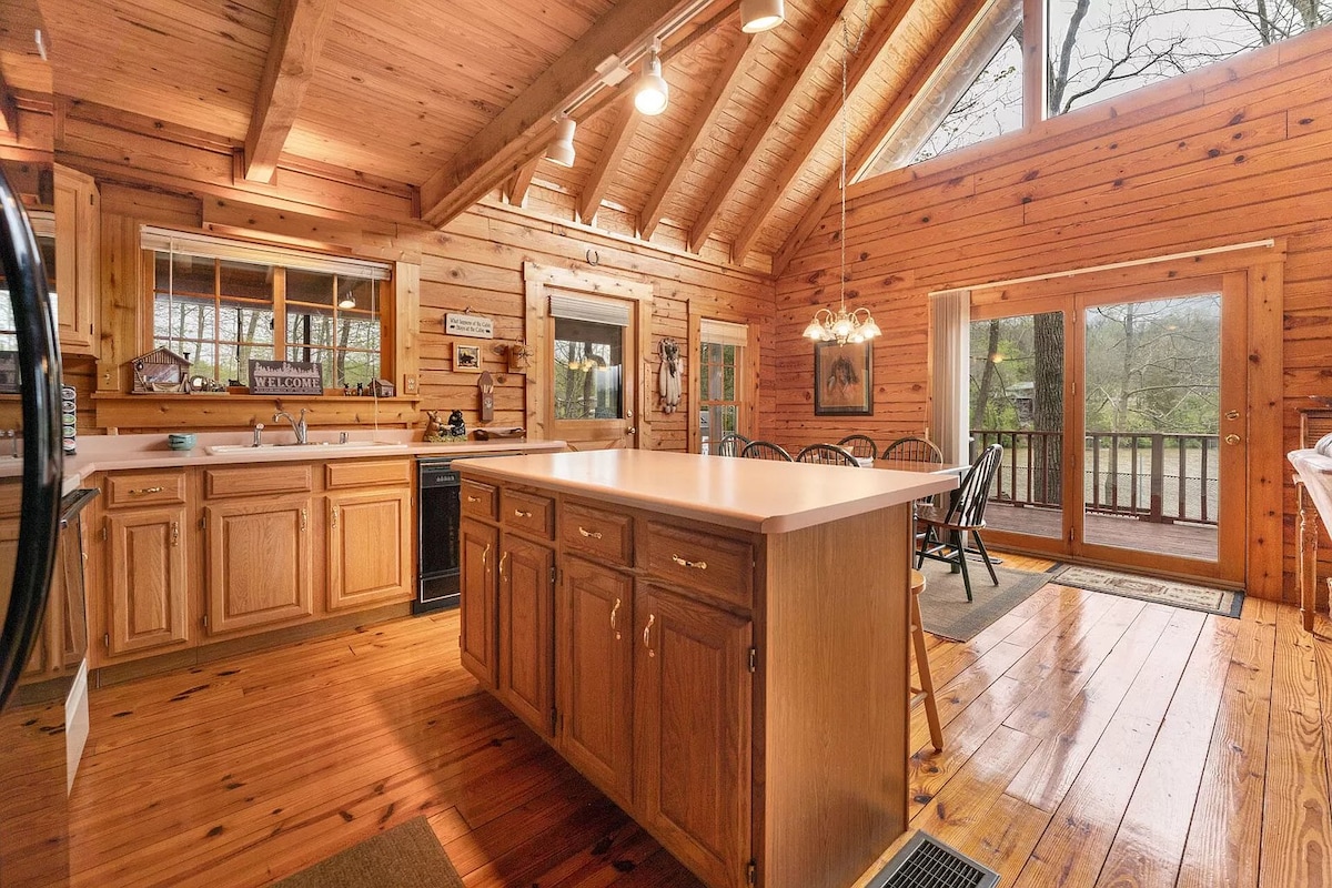 An open kitchen area features wooden cabinetry and a central island, complemented by a spacious dining area with wooden chairs. Large windows invite natural light and provide views of the outdoor surroundings, while the ceiling showcases a dramatic vaulted design with exposed beams.