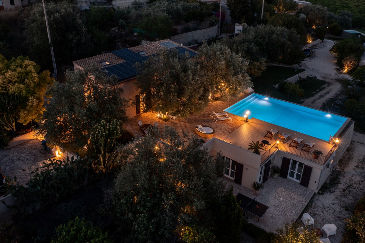 An elevated view of the property showcases a beautifully illuminated infinity pool, surrounded by neatly arranged outdoor seating. The landscape features well-maintained olive trees and gardens, with soft lighting enhancing the pathways leading to the main building.