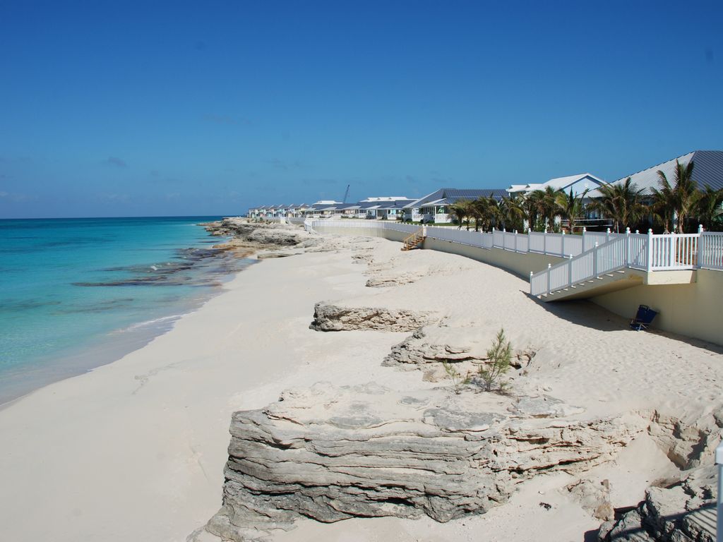 A serene sandy beach stretches alongside inviting turquoise waters, framed by gentle rock formations. A path leads from the shore to modern buildings set in a line, accompanied by palm trees. The clear blue sky enhances the tranquil coastal scene.