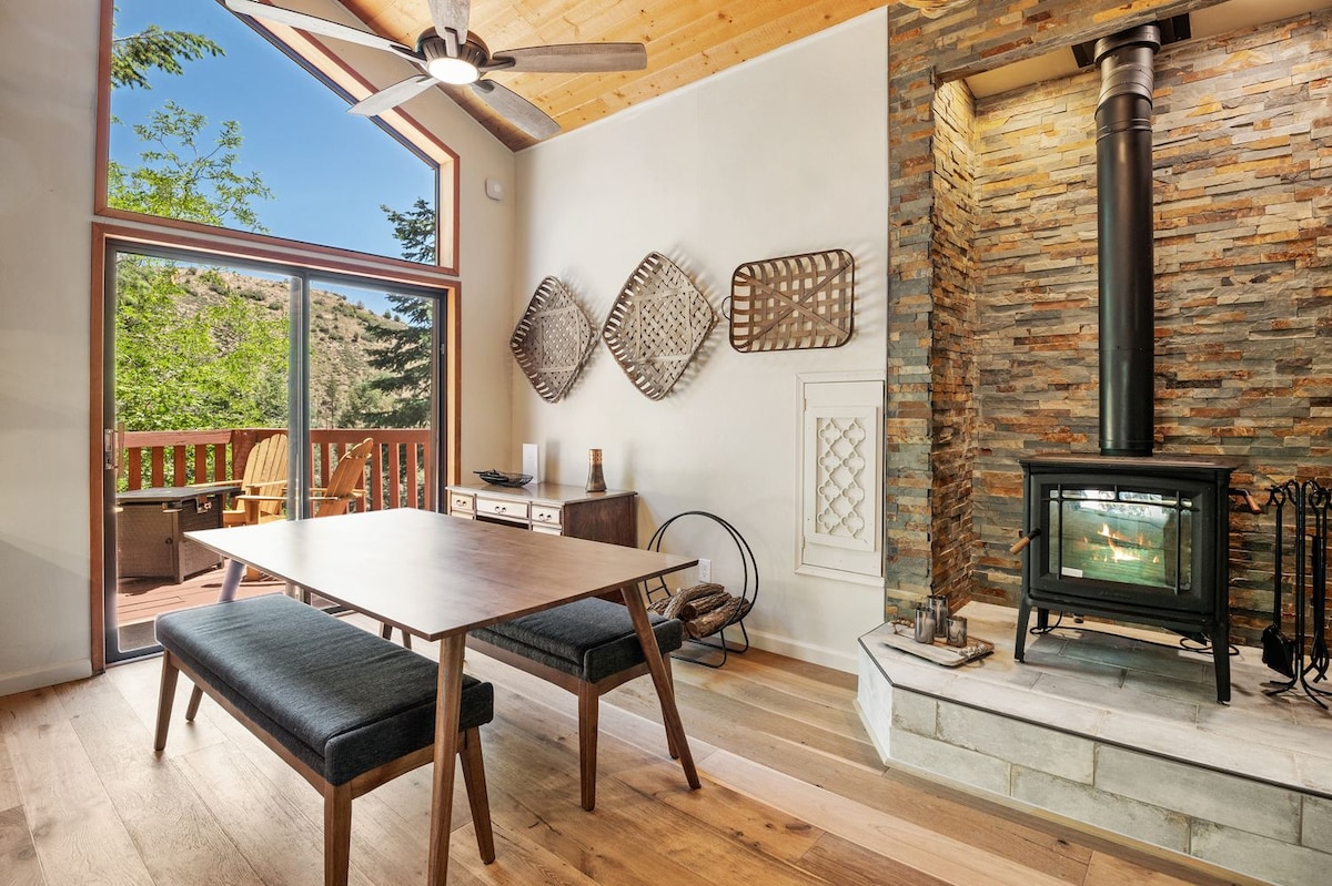A dining area is positioned adjacent to large windows, offering views of the surrounding trees. A wooden table with dark seating sits on polished wood flooring. A stone fireplace serves as a focal point, creating an inviting ambiance with its contemporary design.