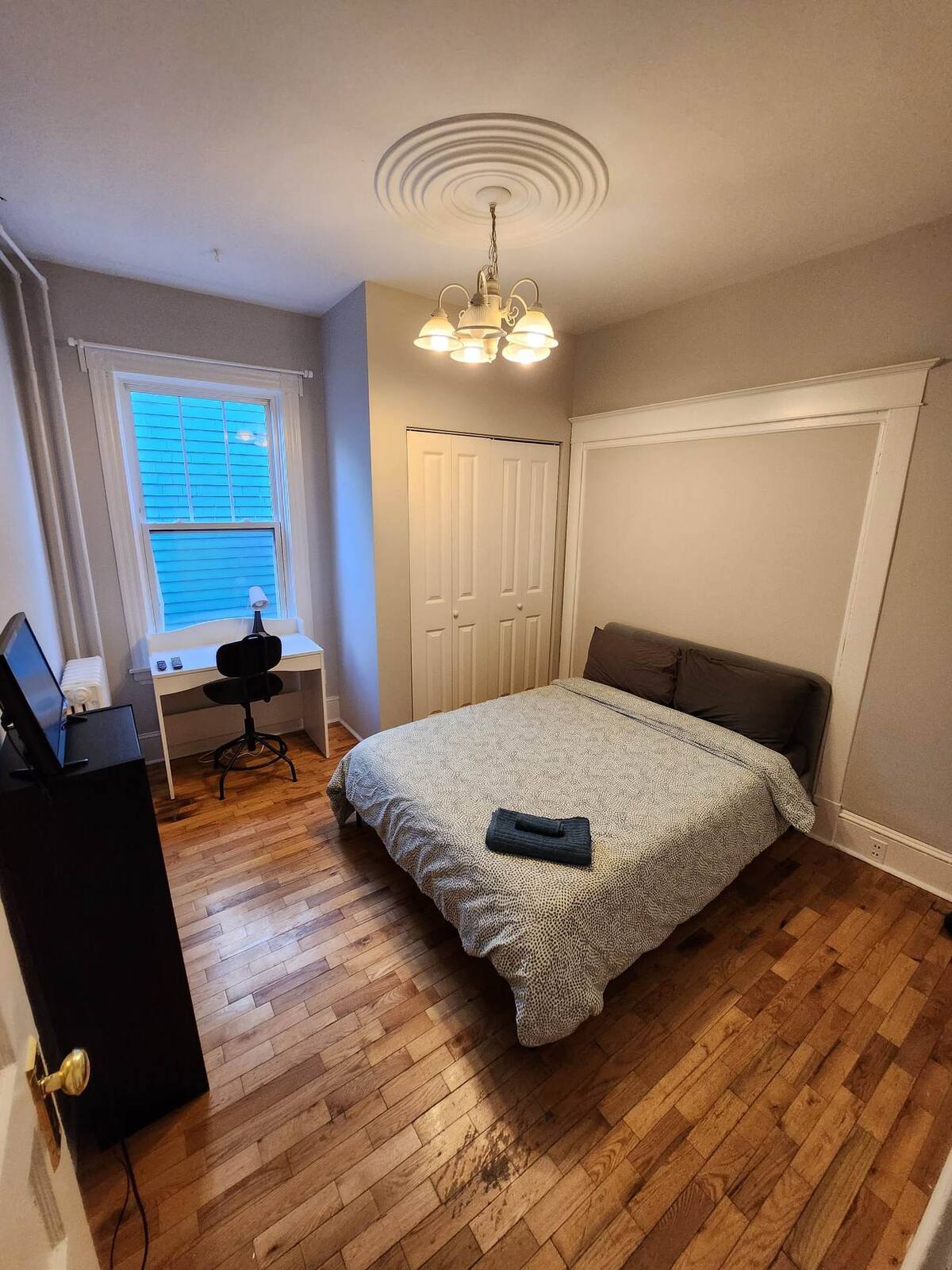 A cozy bedroom features a spacious bed with a patterned coverlet and two pillows. A desk and chair are positioned adjacent to a window with natural light. A closet with sliding doors is visible, and warm wood flooring enhances the room's appeal.