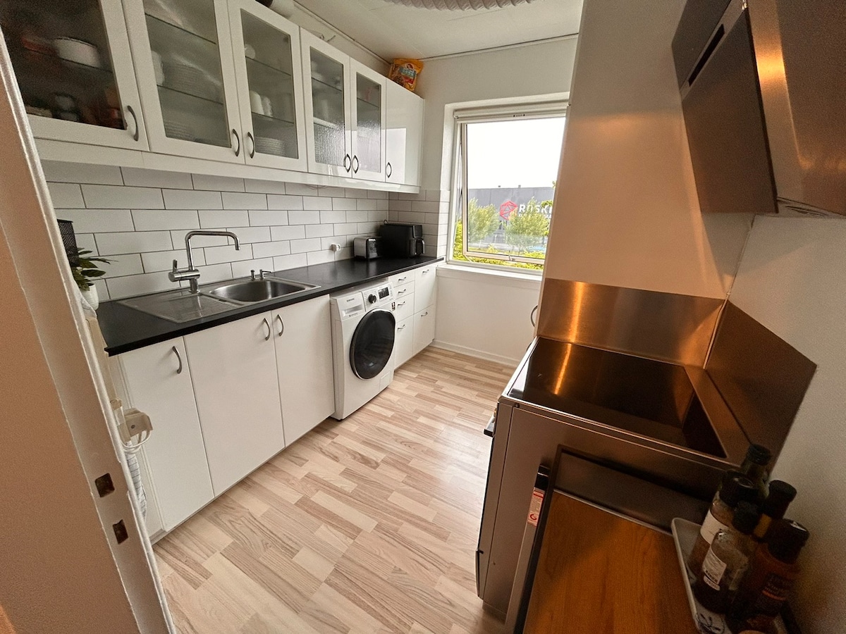 A modern kitchen is shown with white cabinets and a black countertop. The space includes a washing machine, a sink, and stainless steel appliances. Natural light brightens the room through a window, which provides a view of the surroundings.