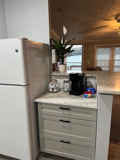 A compact kitchen area is revealed, featuring a white refrigerator and a countertop made of light-colored stone. A small coffee maker sits beside jars and a blue container, with a potted plant adding a touch of greenery. Natural light filters in from a nearby window.