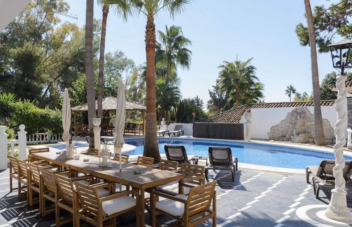 An outdoor dining area features a large wooden table surrounded by comfortable chairs, inviting gatherings under the sun. The nearby swimming pool is framed by sun loungers and tropical landscaping, creating a relaxing atmosphere in a well-maintained garden.