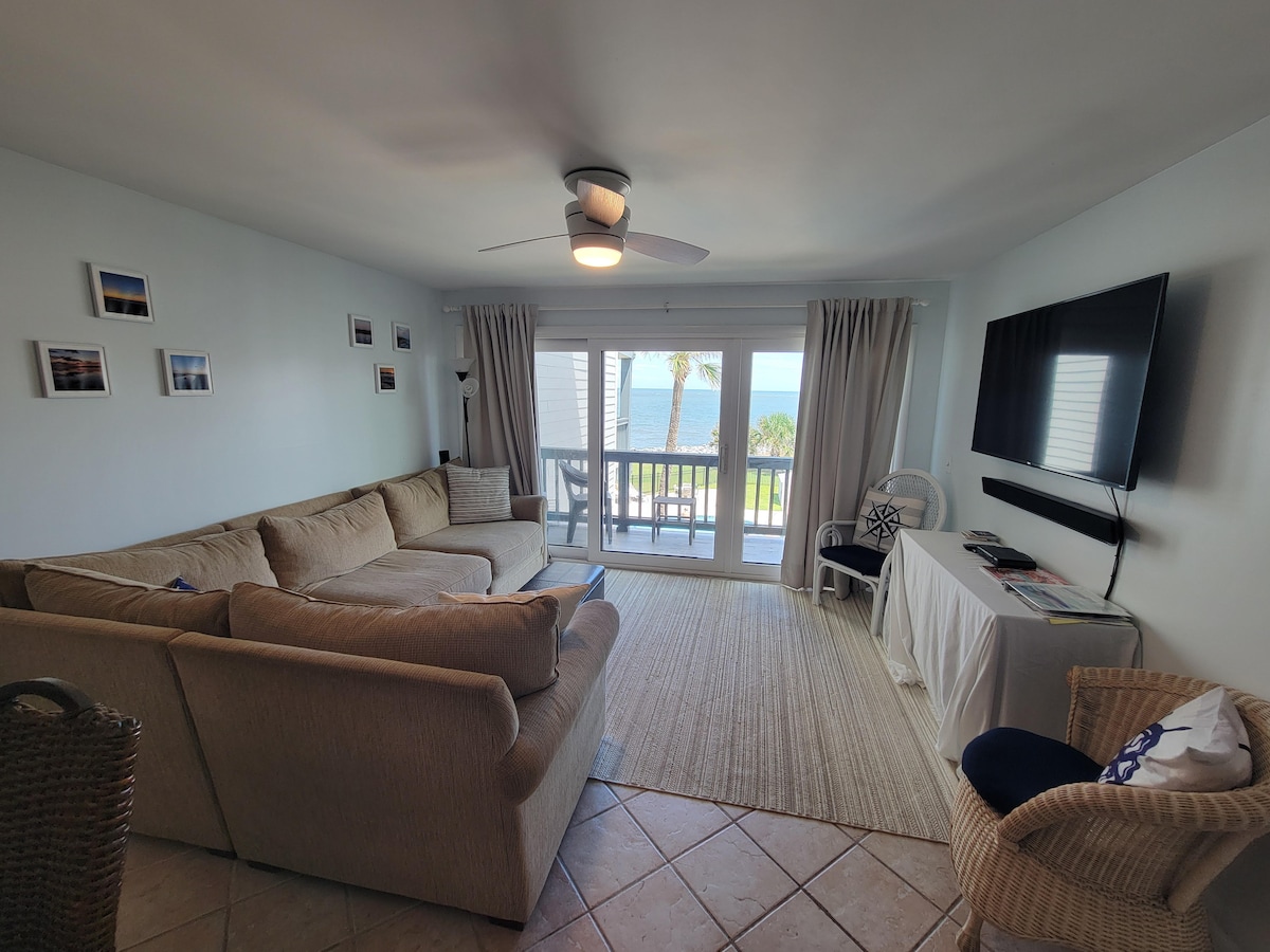 A spacious living area is highlighted by a large sectional sofa, providing ample seating. Light-colored walls and soft curtains allow natural light to illuminate the space. A TV is mounted on the wall opposite the sofa, with a view of the ocean visible through sliding glass doors.