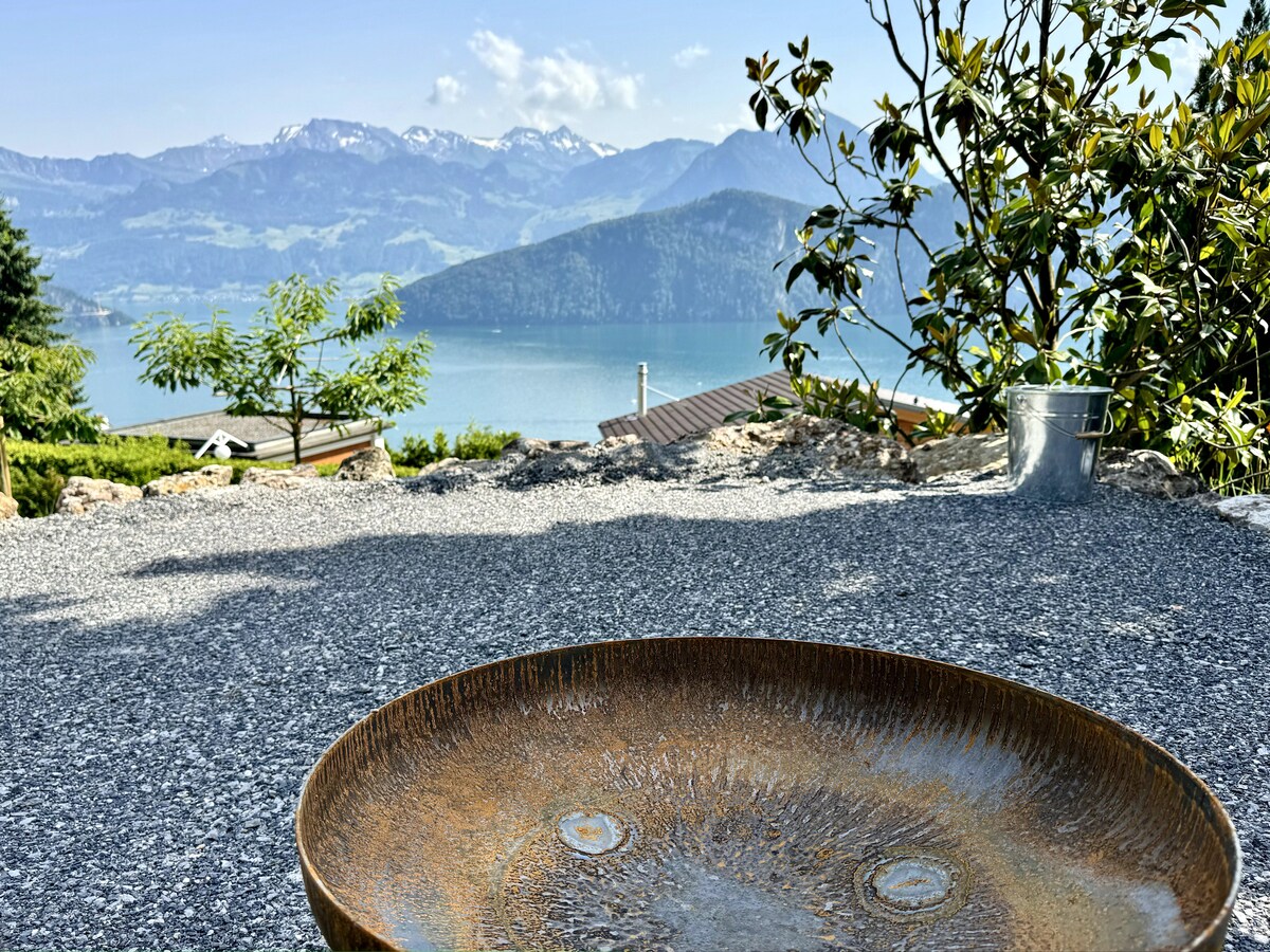 A shallow, rustic bowl is centered in the foreground, surrounded by gravel. In the background, a scenic view of a calm lake and majestic mountains is visible under a clear sky, providing a serene natural backdrop.