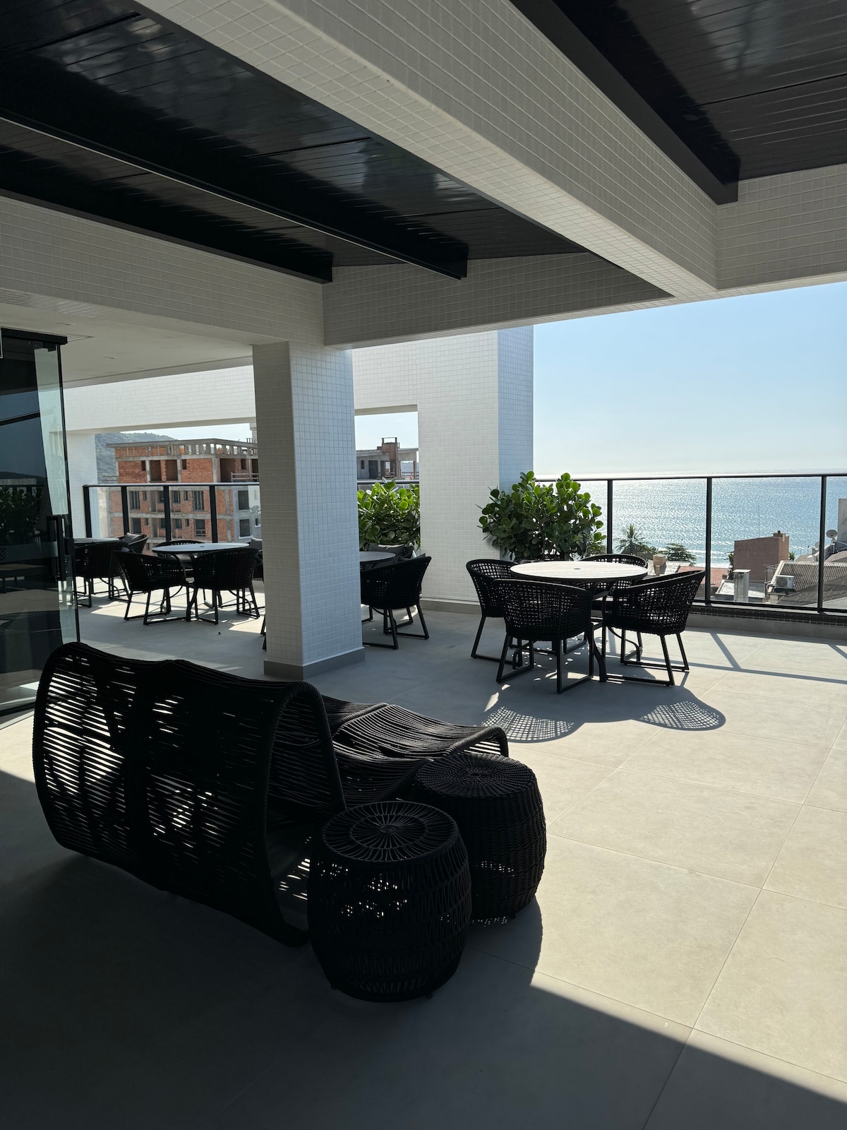 An open terrace area is presented, featuring modern furniture in dark tones. A circular dining table with surrounding chairs sits beside comfortable lounge chairs. Lush greenery is visible along the edges, with an unobstructed view of the sea in the background under bright, clear skies.