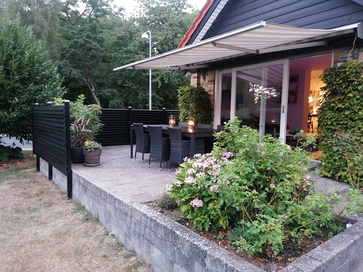 An outdoor terrace is featured, providing a spacious dining area with a large table and several chairs. Potted plants and blooming flowers add greenery, while an awning provides shade. Soft lighting from candles creates a welcoming ambiance in the evening.
