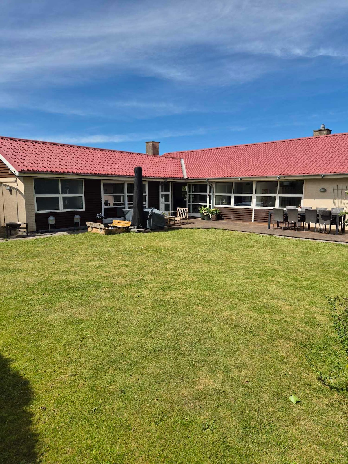 A spacious backyard is visible, featuring well-maintained green grass under a clear blue sky. Outdoor seating areas are arranged, with lounge furniture positioned near a tall, modern patio heater. The house, with a red roof and large windows, provides an inviting backdrop.