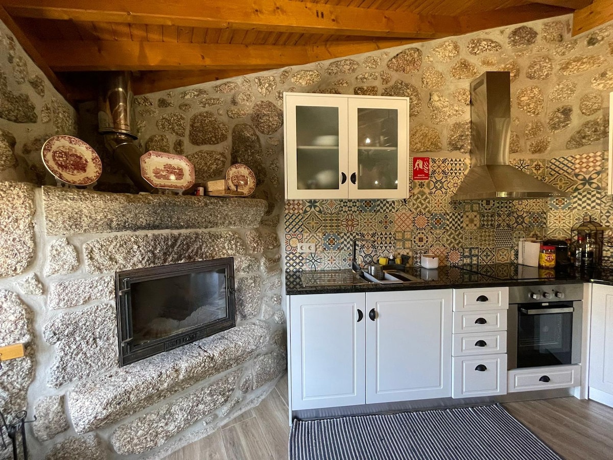 The kitchen features stone walls complemented by decorative tiles and wooden beams. A built-in stone fireplace is positioned next to modern appliances, including an oven and a hood. White cabinetry and a striped rug add to the cozy ambiance of this functional space.