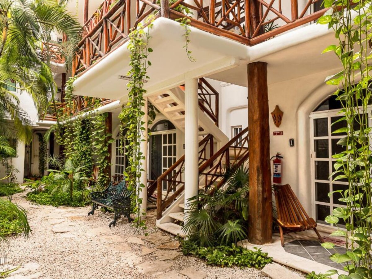 A lush courtyard is surrounded by white walls and features several tropical plants. A staircase leads to the upper level, with a wooden railing and steps. Two seating areas are visible: a dark green bench and a wooden bench, both positioned on a stone pathway.