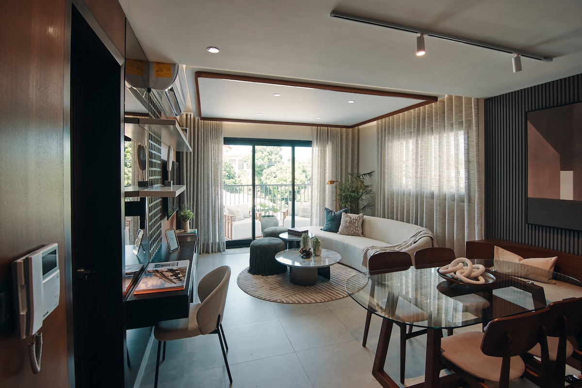 An open-concept living area features a plush sectional sofa, a circular coffee table, and a dining table surrounded by stylish chairs. Large glass doors lead to a balcony, allowing natural light to illuminate the space, complemented by elegant decor and modern furnishings.