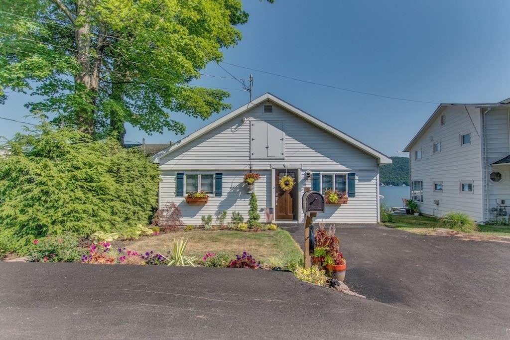 The exterior of the cottage is viewed from the front, featuring a well-maintained lawn with colorful flower beds and a mailbox. Large windows reflect the surrounding greenery and provide a glimpse of Keuka Lake in the background, offering a welcoming entrance to the cozy accommodation.