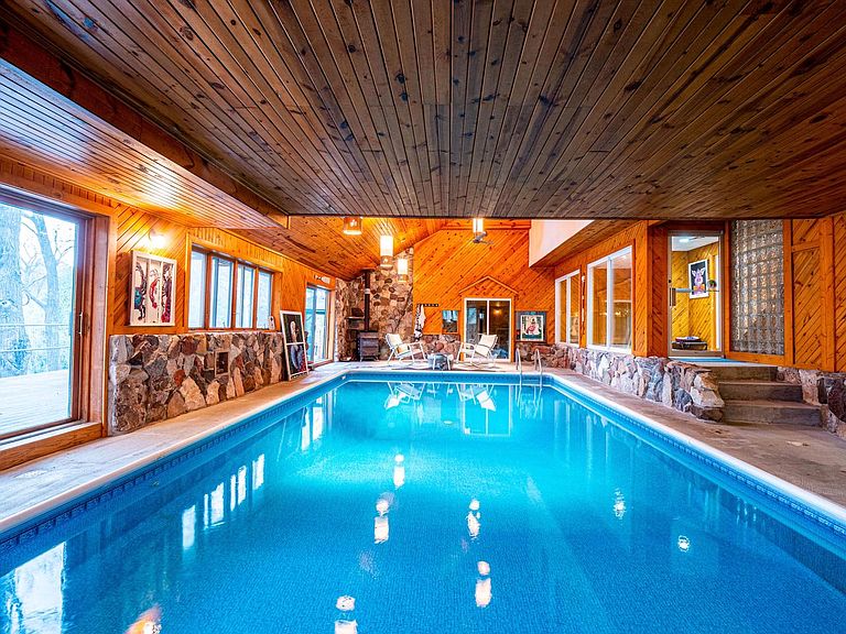An indoor pool is framed by natural stone and wood paneling, with large windows allowing natural light. Lounge chairs are arranged at one end of the pool, offering a relaxed setting. The pool area is spacious, creating a calming atmosphere for leisure activities.