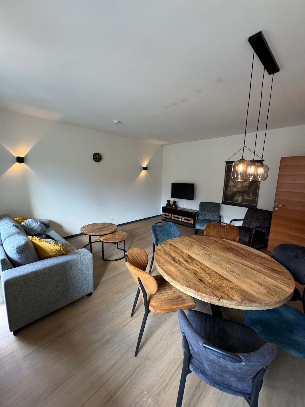 A modern living and dining area is presented with a round wooden table surrounded by several chairs. A sleek gray sofa and armchairs create a comfortable seating arrangement, with soft lighting enhancing the relaxed ambiance. A wall-mounted television is visible, contributing to the space's functionality.