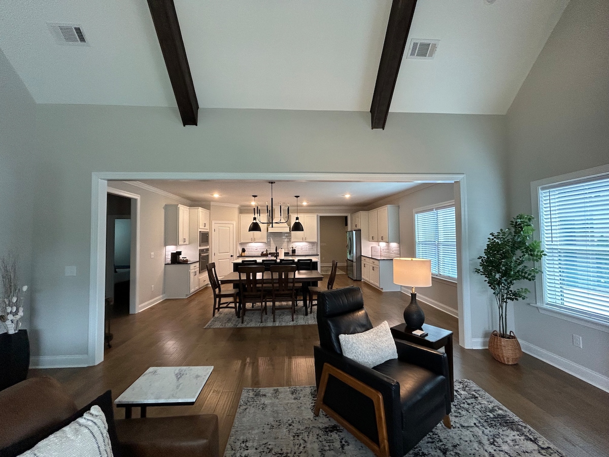 An open living space features vaulted ceilings with dark wood beams. A modern seating area includes a leather armchair beside a side table with a lamp, while a spacious dining area is visible in the background, showcasing a large table surrounded by eight chairs and a fully equipped kitchen.