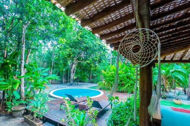 A tranquil outdoor area features a circular swimming pool surrounded by lush greenery. Wooden decking holds comfortable lounge chairs, and a decorative dreamcatcher hangs from the rustic structure, adding a serene touch to the natural setting.