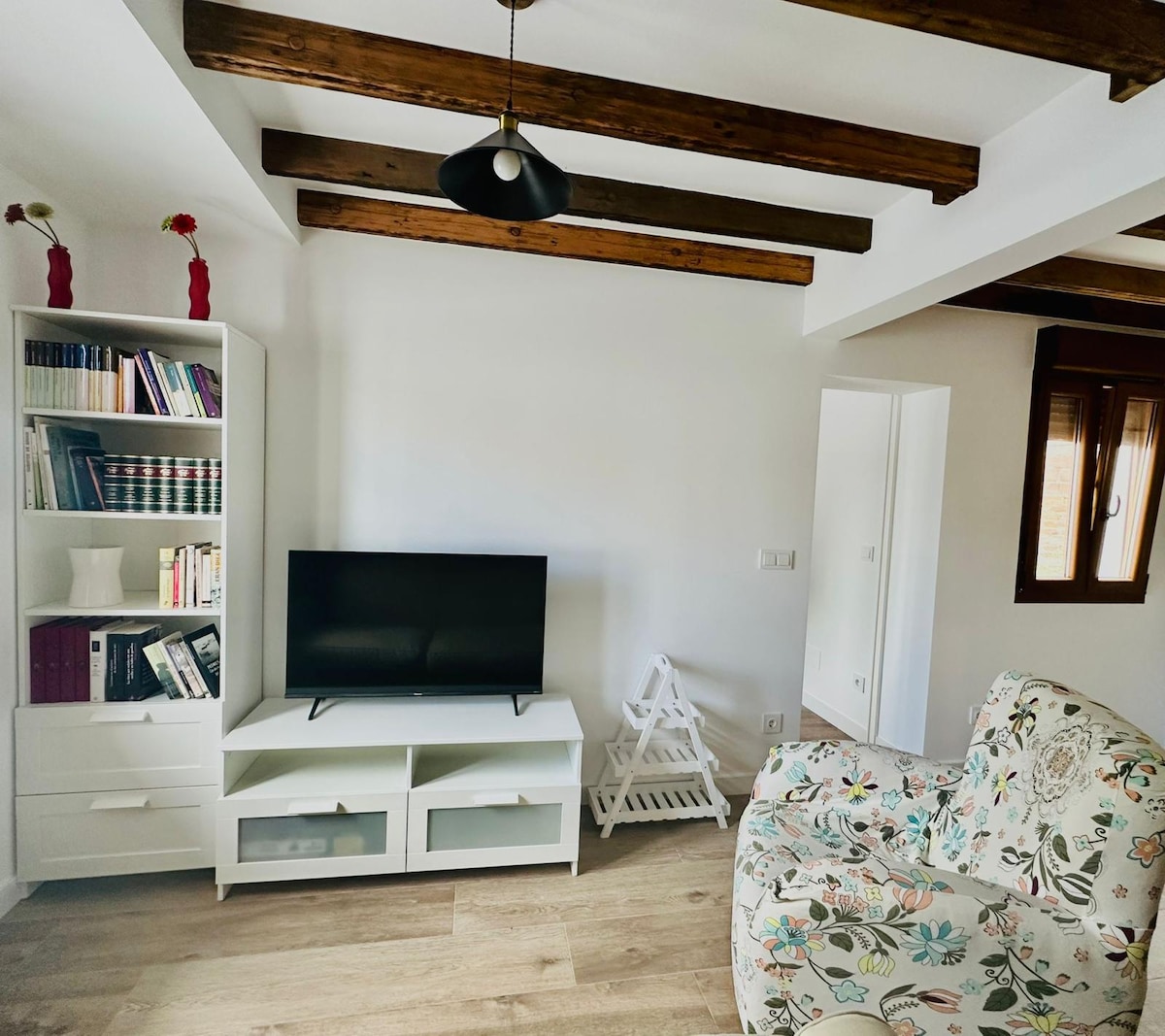 A bright living area is presented, featuring a white entertainment unit that holds a flat-screen television. A floral-patterned armchair adds a pop of color, while wooden beams adorn the ceiling. A bookshelf is filled with books and a couple of decorative items, enhancing the inviting space.
