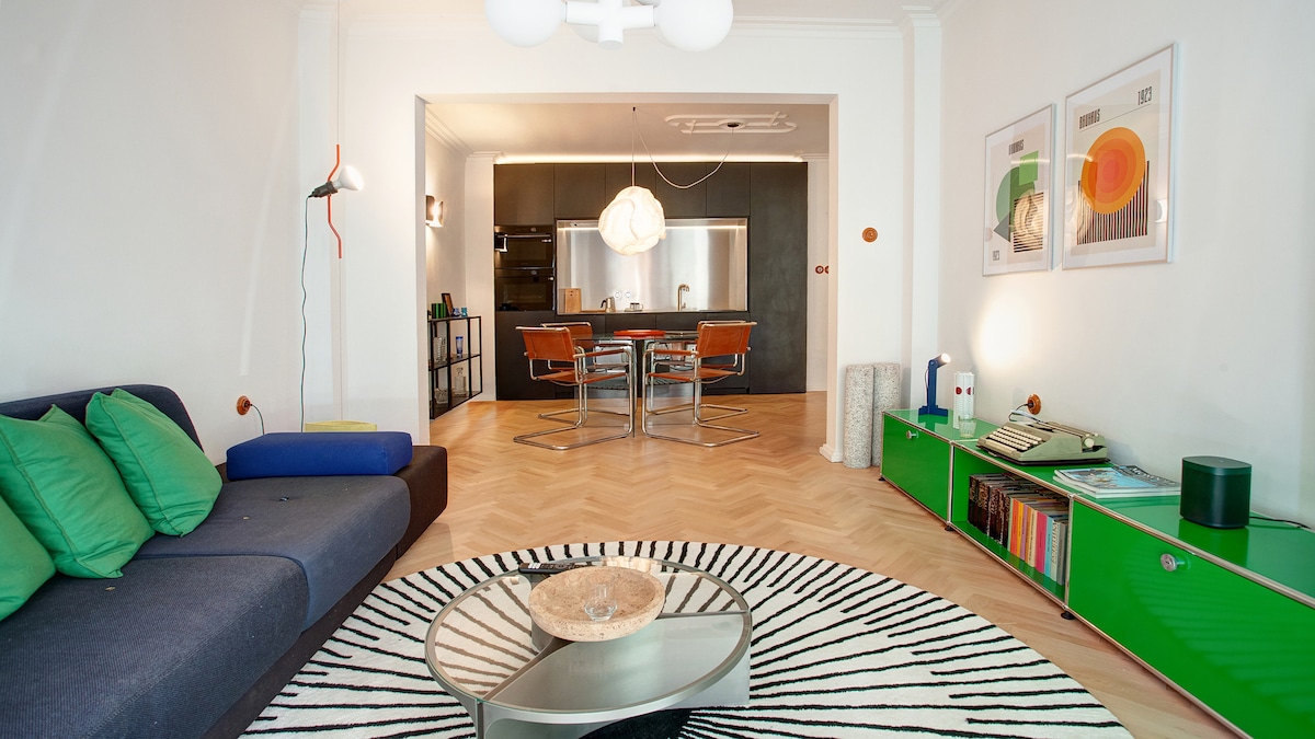 A spacious living area features a blue sofa adorned with green accent pillows and a patterned rug. A round coffee table sits at the center, surrounded by a retro-style dining area visible in the background, complete with modern chairs and a sleek kitchen.