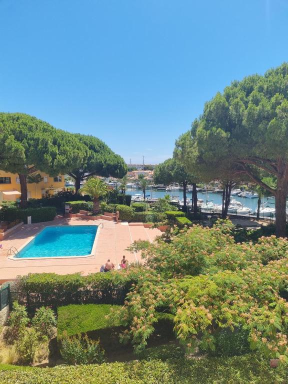 A view of a pool surrounded by landscaped gardens and mature trees in a residential complex. In the background, a marina with moored boats is visible under a clear blue sky, enhancing the serene outdoor atmosphere.