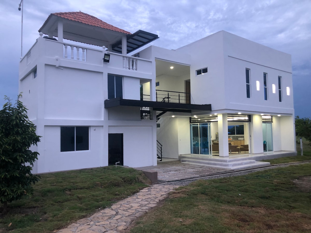 A modern villa showcases a minimalist design with a light-colored exterior. It features multiple levels, large windows for natural light, and a pathway leading to the entrance. A spacious balcony is visible on the upper floor, overlooking the surrounding green landscape.