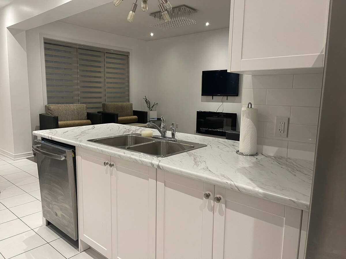 A modern kitchen features a marble countertop with a double sink, complemented by a sleek stainless-steel dishwasher. Soft lighting highlights the contemporary cabinetry. A seating area with two armchairs is visible in the background, along with a television mounted on the wall.