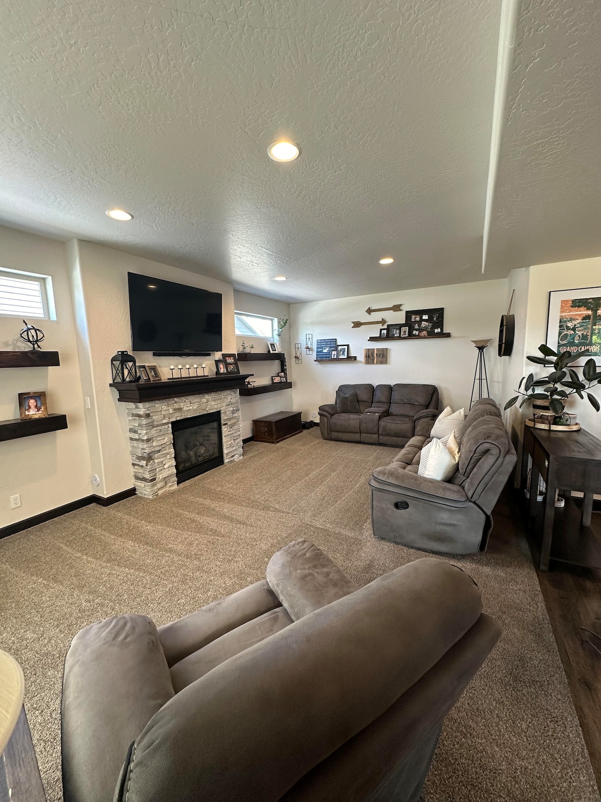 A comfortable living area is highlighted by a stone fireplace and a mounted flat-screen TV. Plush seating is provided by two reclining sofas and a single armchair. Natural light filters through small windows, and decorative shelving adds character to the space.