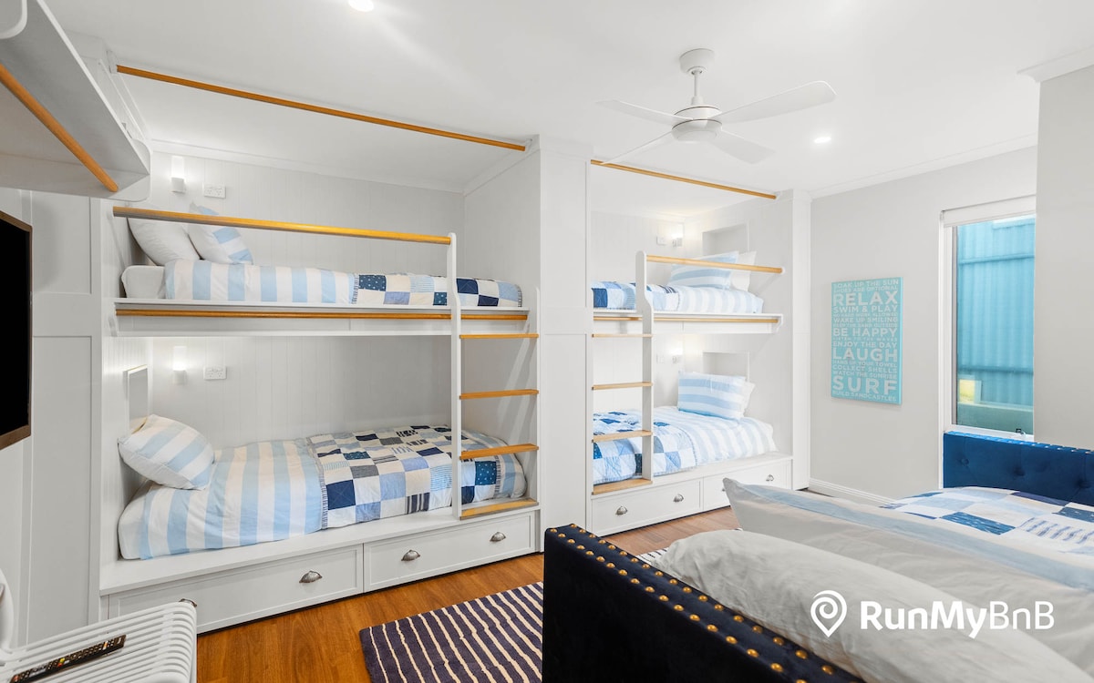 Ugodna soba s krevetima na kat s posteljinom u bijelo-plave pruge i s više kreveta na kat s drvenim okvirom. Soba je čista i prozračna, ima stropni ventilator, a na zidu je motivacijski natpis na kojem piše „RELAX BE HAPPY LAUGH SURF.”