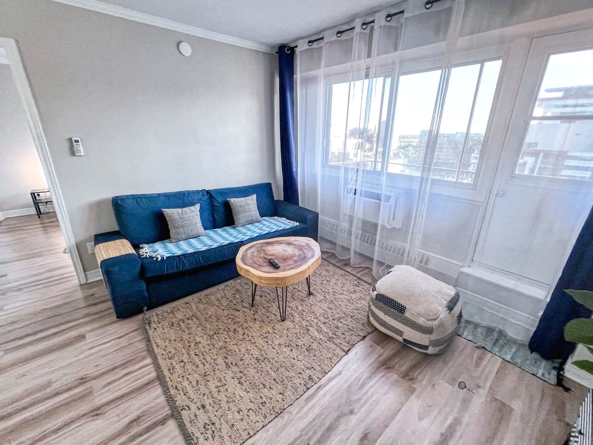 A bright living area features a comfortable blue sofa adorned with two cushions, positioned next to a round wooden coffee table. Sheer curtains frame large windows, allowing natural light to illuminate the space. A woven pouf and a decorative area rug add texture to the room.