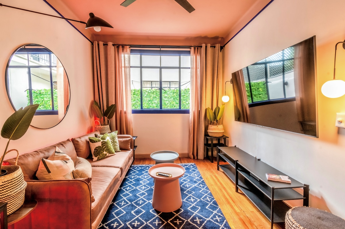 The living area features a cozy sofa adorned with patterned pillows. A modern coffee table is positioned on a decorative rug. Large windows allow natural light to illuminate the space, complemented by indoor plants and contemporary lighting fixtures.