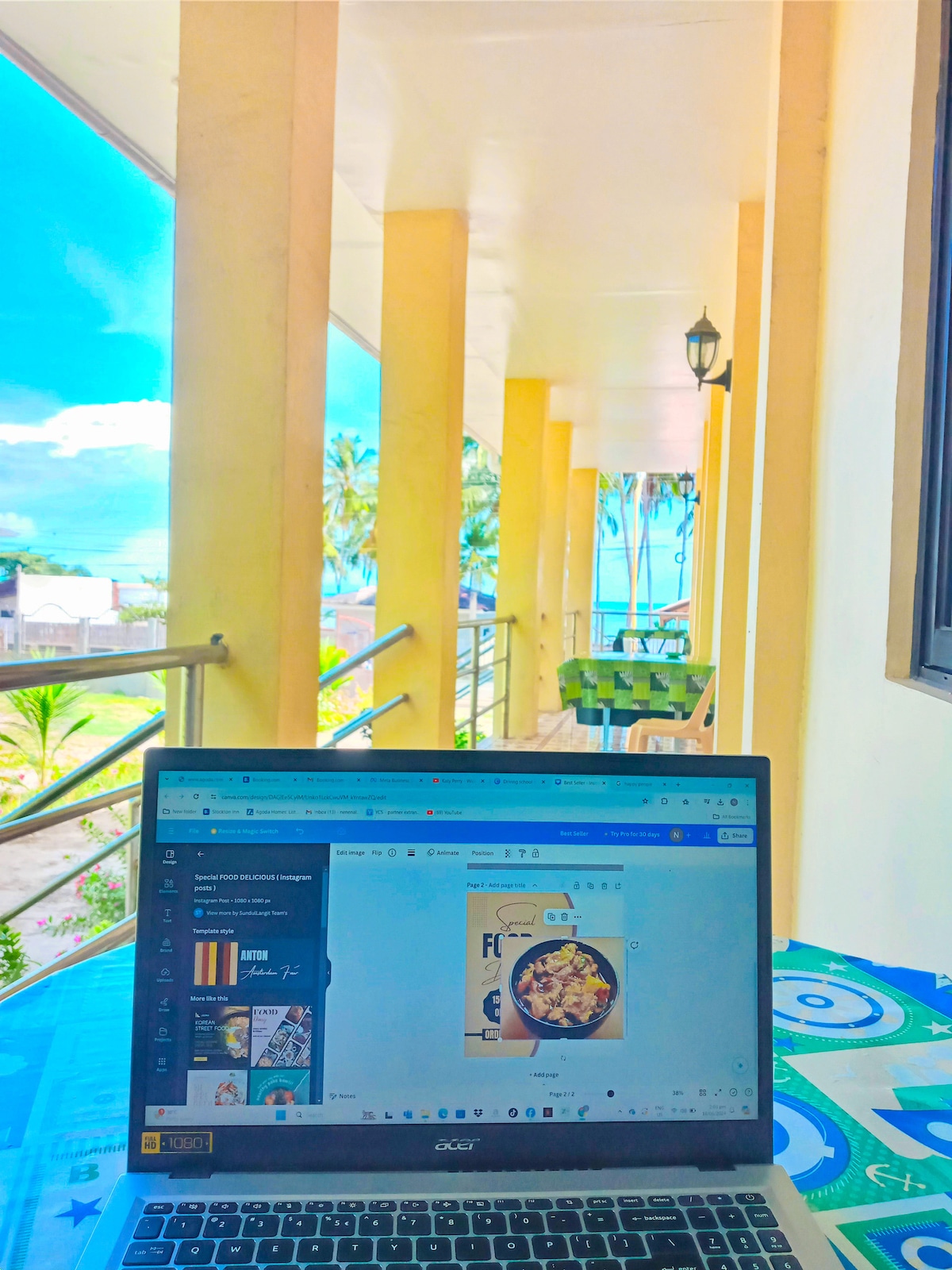 An outdoor workspace is shown with a laptop open on a table, facing a vibrant view of palm trees and the sea. Soft sunlight illuminates the area, and a charming yellow column supports the covered terrace. Green seating can be seen in the background.