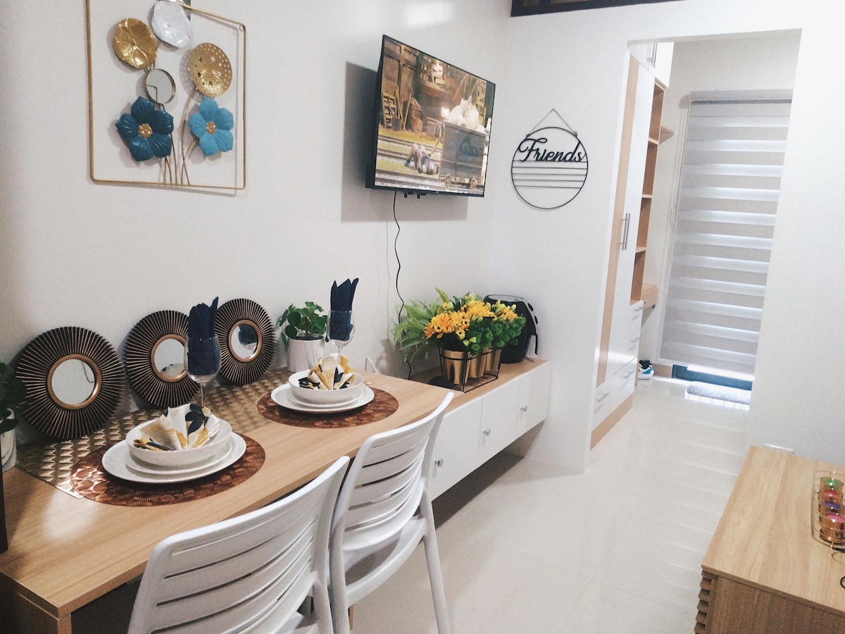 A dining area features a light wooden table set for two, with elegant dinnerware and decorative centerpieces. A wall-mounted television displays a show, while framed art and decorative plates adorn the wall. A bright arrangement of yellow flowers adds a touch of color.